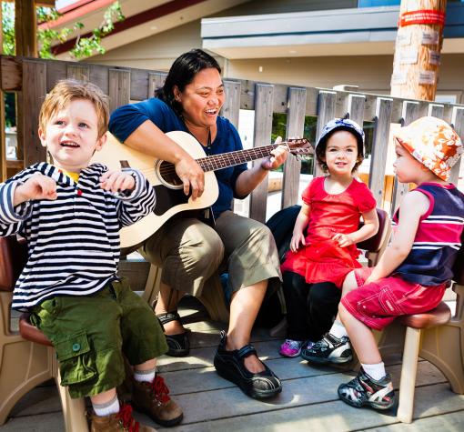 A woman with dark hair is sitting on a patio outside and playing a guitar. She is smiling at three 3-year-old children sitting around her in summer clothes.