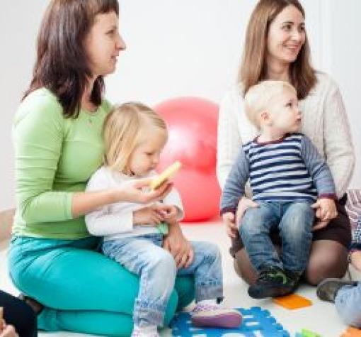 A group of young moms are sitting in a play room with their 3 year old children on their laps. They are smiling and listening to a speaker who is off screen.